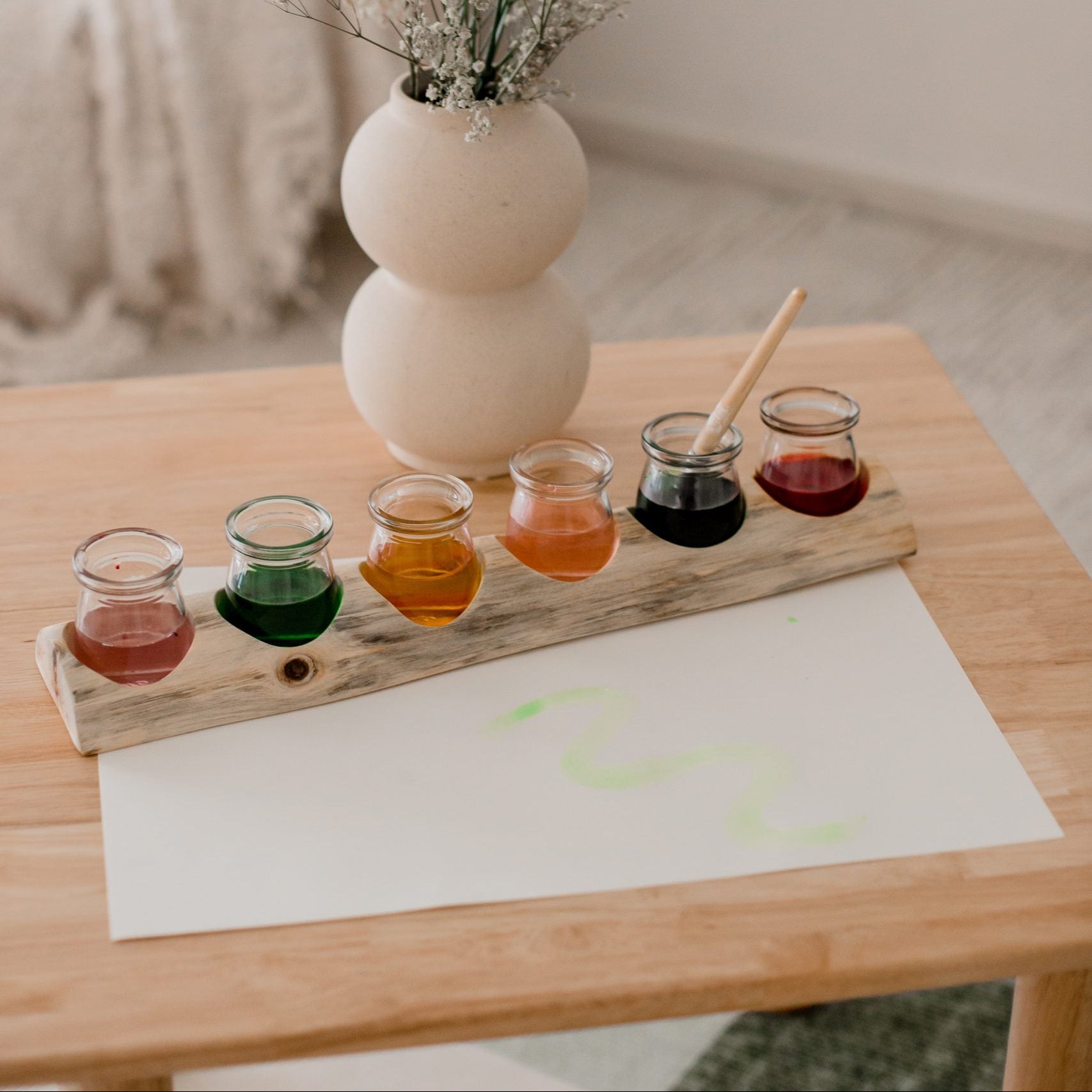 Set of small glass containers with colorful liquids on a wooden board, placed on a table with a vase and flowers in the background.