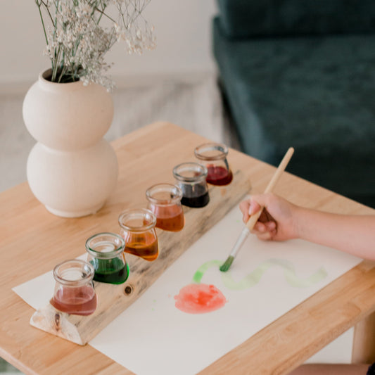 Person painting a flower on paper with watercolor paints on a wooden table.