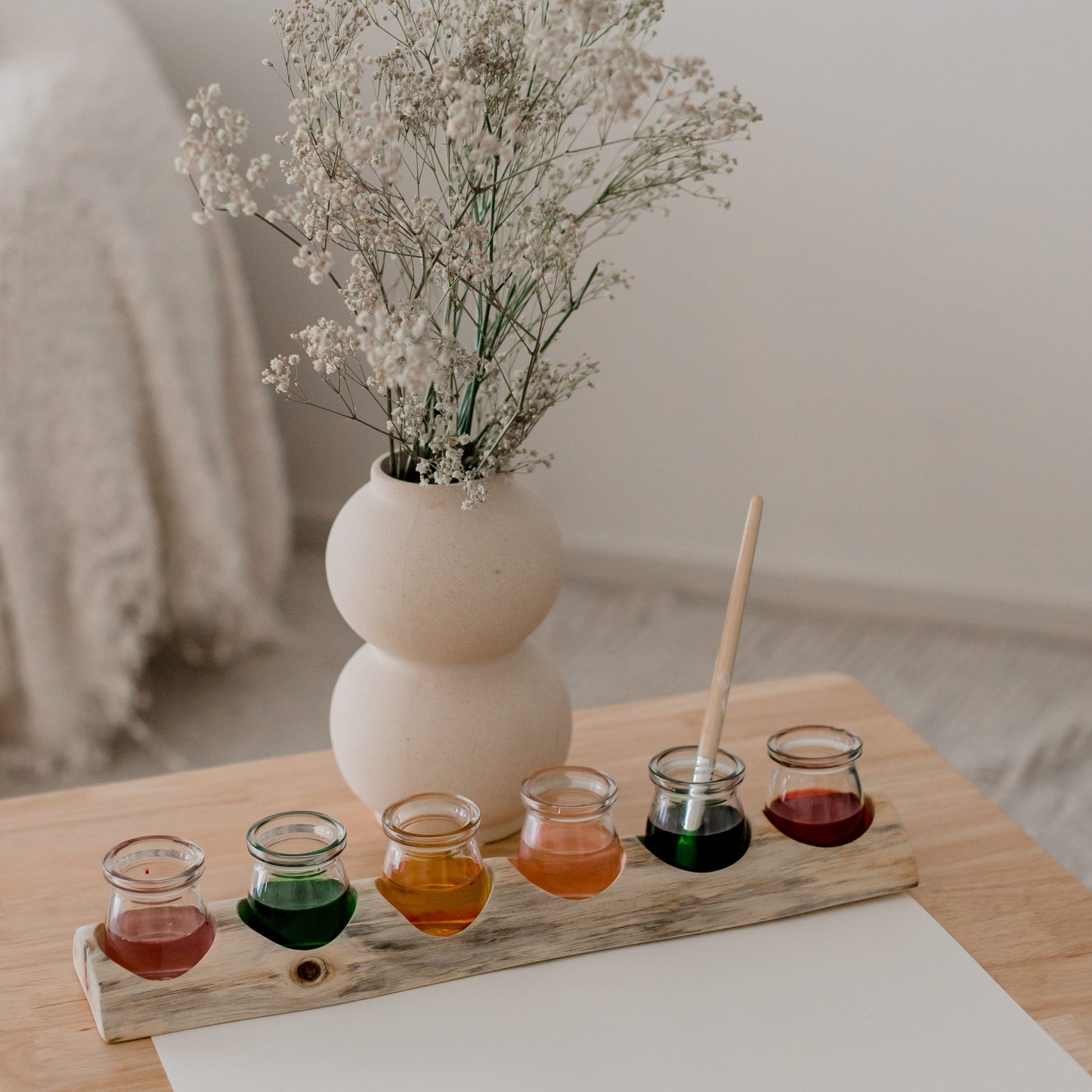 Set of small glass containers with colorful liquids on a wooden board, accompanied by a vase with dried flowers.